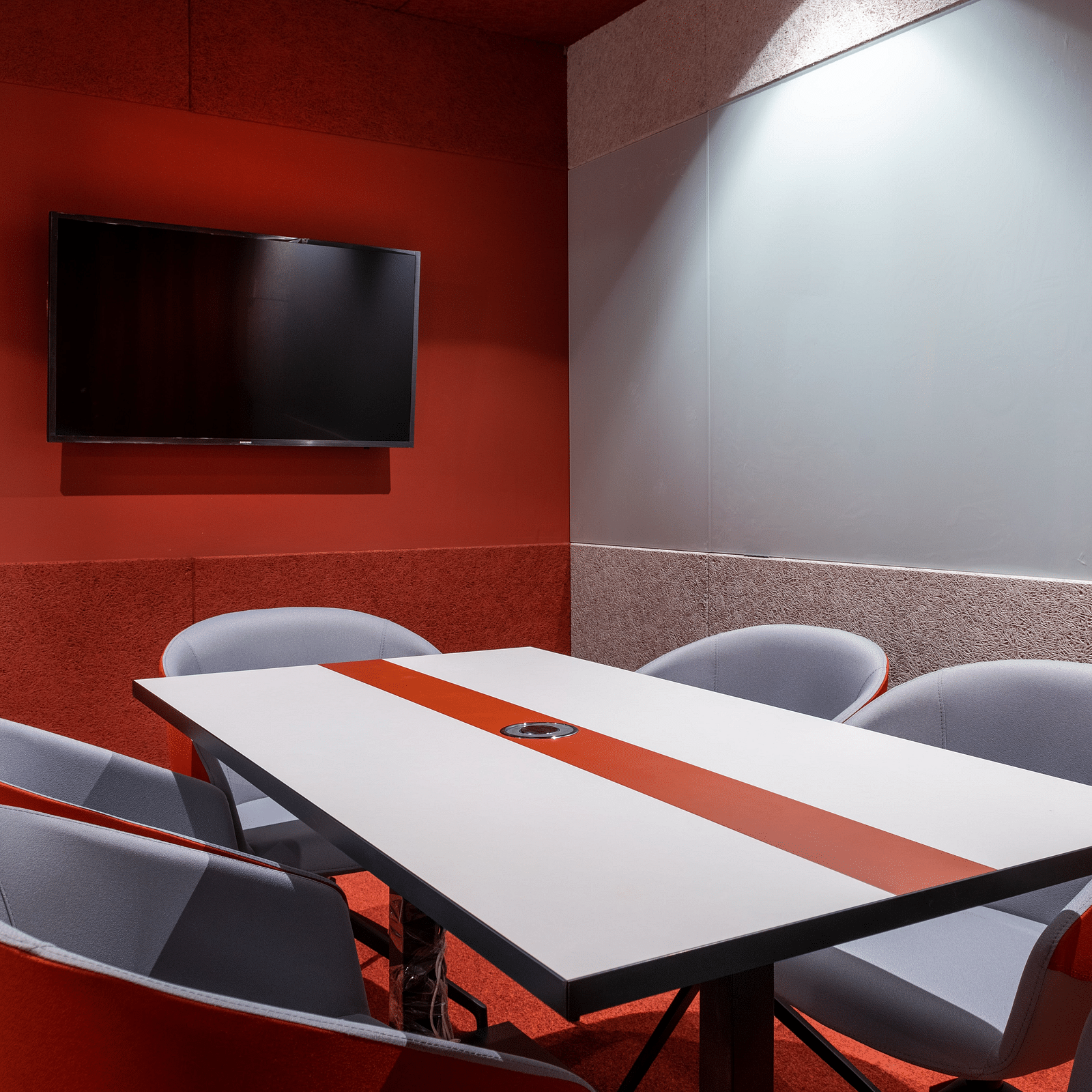 A modern meeting room with a red and white theme, featuring a long white table with a red stripe, surrounded by curved red chairs and a TV mounted on the wall