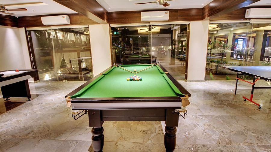 A green billiards table ready for play, with cues and balls set, in a spacious room with wooden beams and glass walls offering a view into an adjacent area