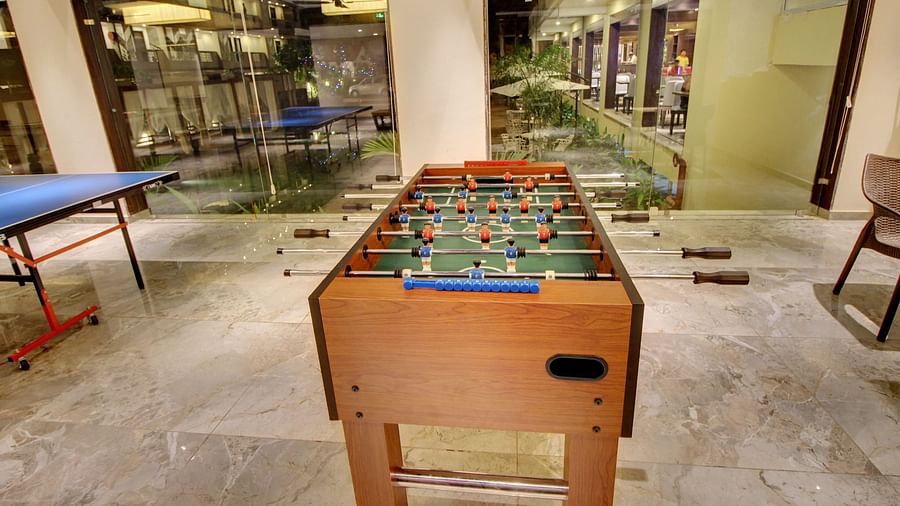 Foosball table in focus with game handles and miniature players, in a leisure room with other gaming tables and a view of the outdoor area through glass walls