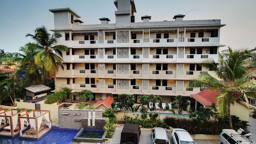 Exterior of a multi-story building with intricate balcony designs, overlooking a swimming pool and surrounded by tropical trees and parked cars