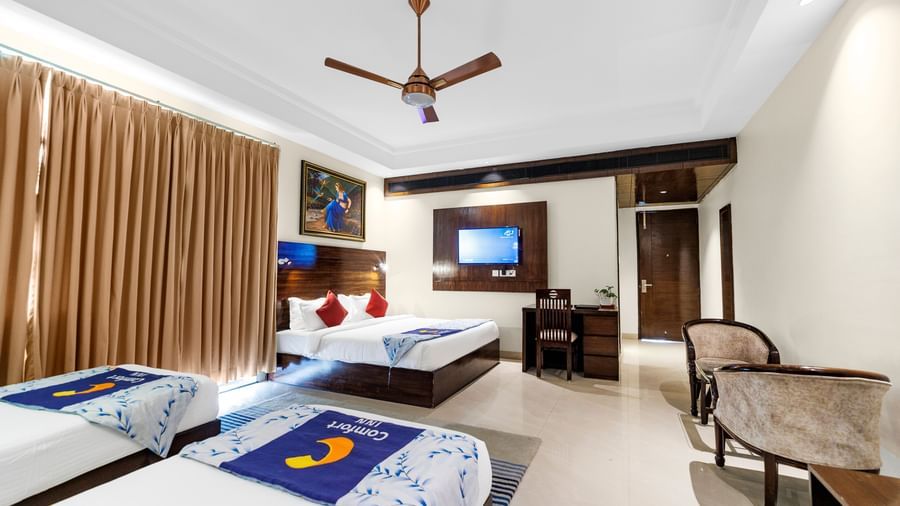 A wide room with a king size bed and twin beds featuring full-length curtains, ceiling fan, desk with chair, and wall-mounted TV at Inde Reva Resorts Rishhikesh.