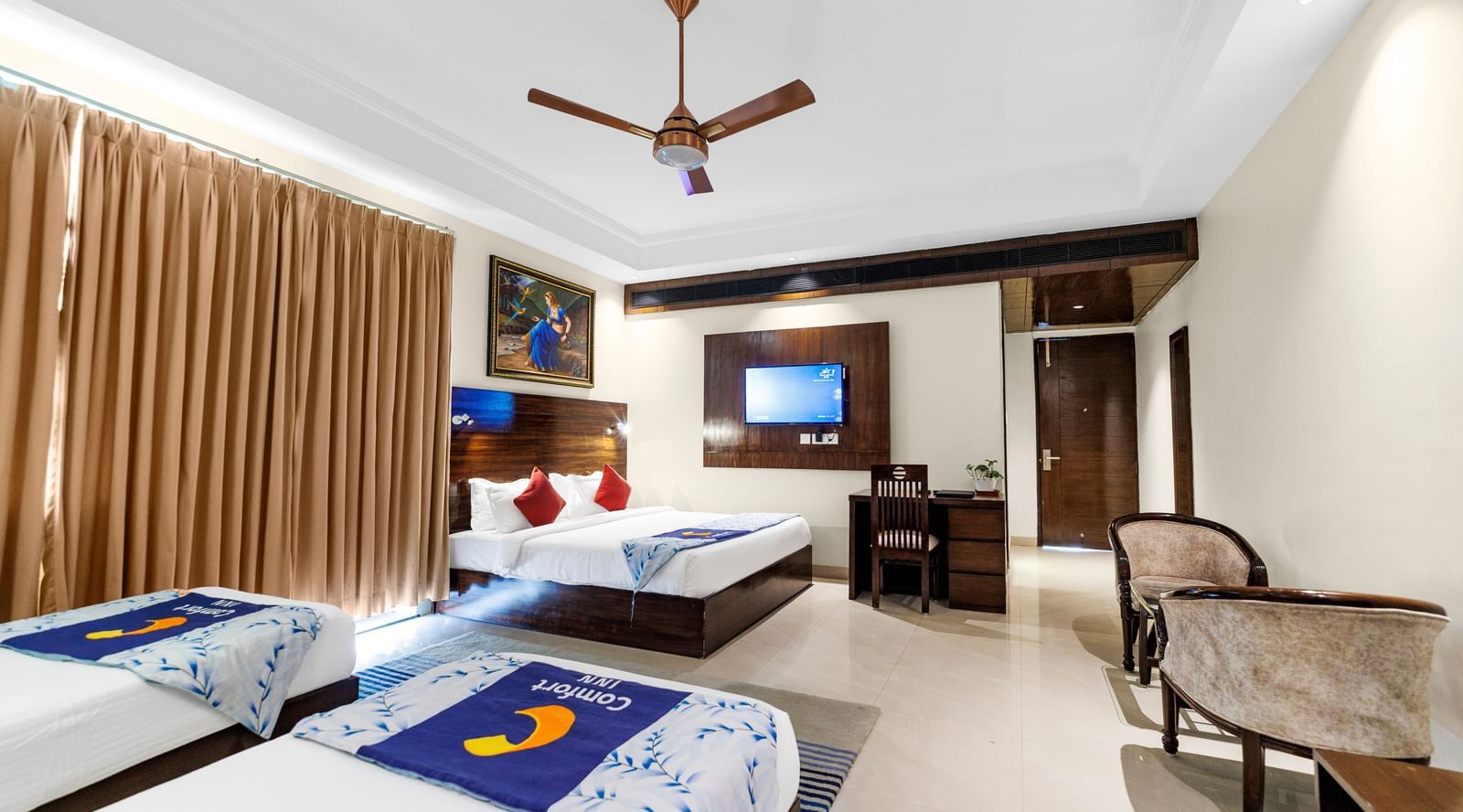A wide room with a king size bed and twin beds featuring full-length curtains, ceiling fan, desk with chair, and wall-mounted TV at Inde Reva Resorts Rishhikesh.