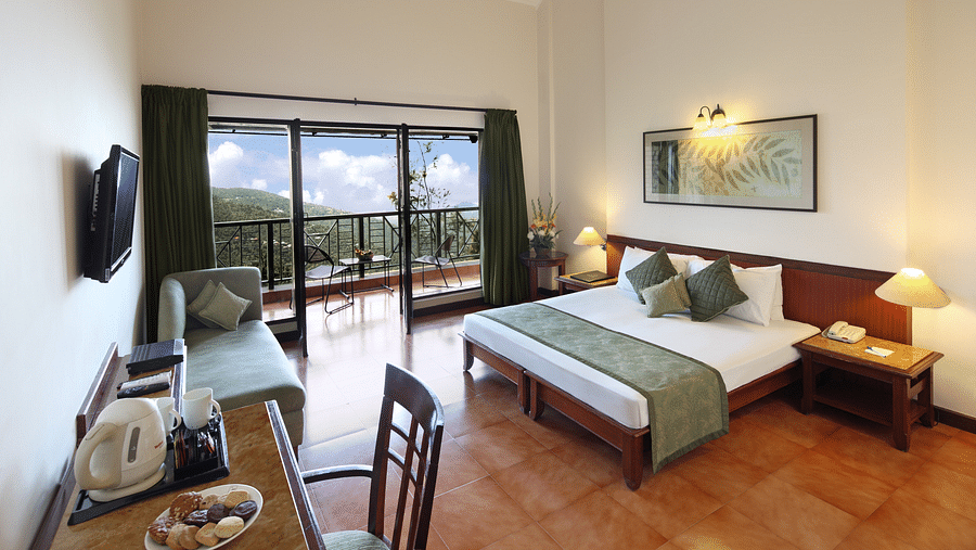 A bedroom featuring a study table and chair, a bedside table with a lamp, and a balcony offering a stunning mountain view -  Baikunth resort, kasauli 
