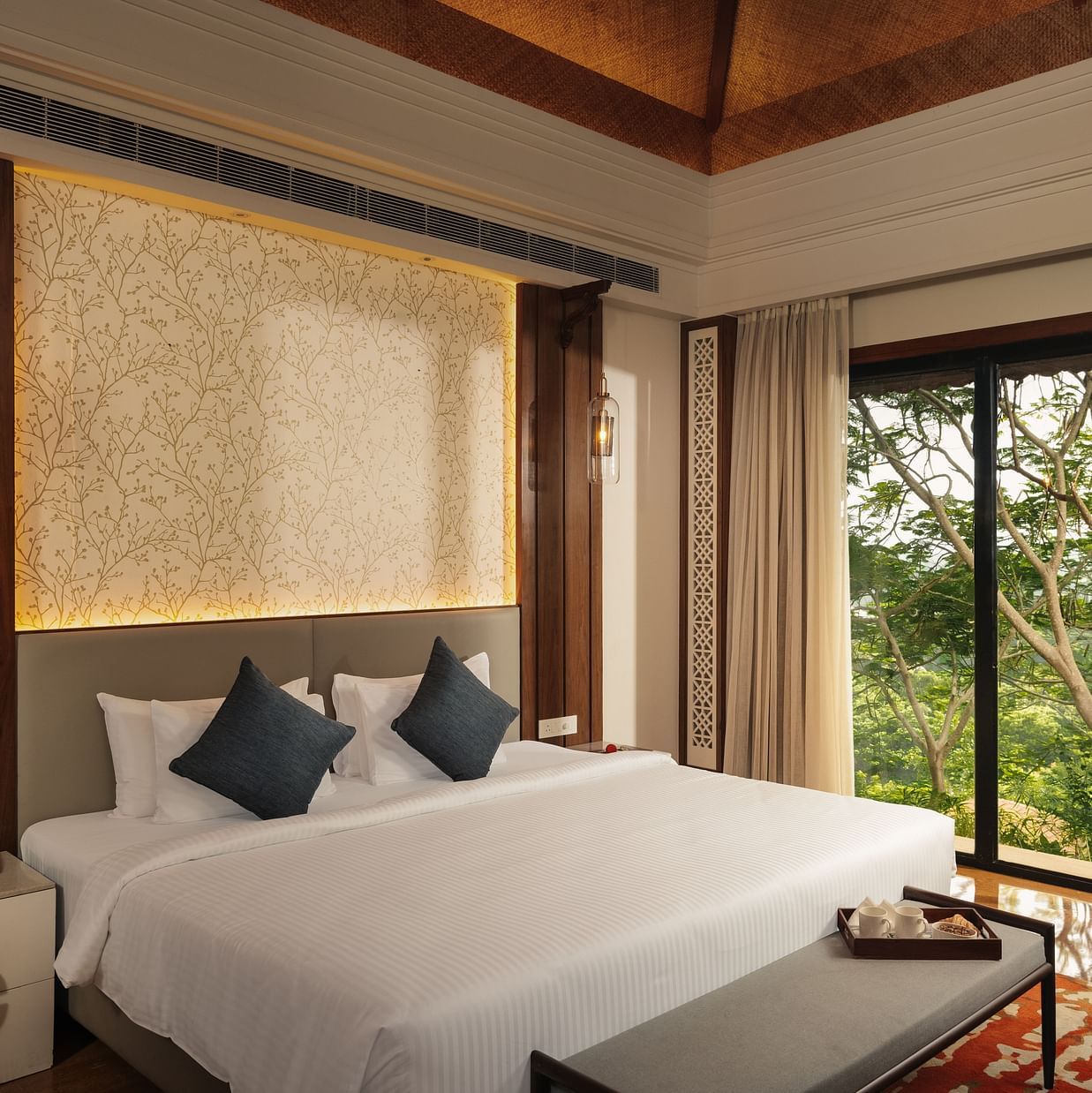 A well-appointed hotel bedroom at The Ananta Udaipur. A large bed with white linens and dark cushions is the focal point. A bedside table with a lamp sits to the left. Large windows offer a view of lush greenery outside. A wooden bench is placed at the foo