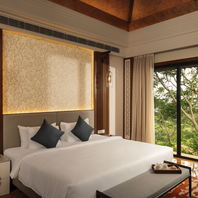 A well-appointed hotel bedroom at The Ananta Udaipur. A large bed with white linens and dark cushions is the focal point. A bedside table with a lamp sits to the left. Large windows offer a view of lush greenery outside. A wooden bench is placed at the foo