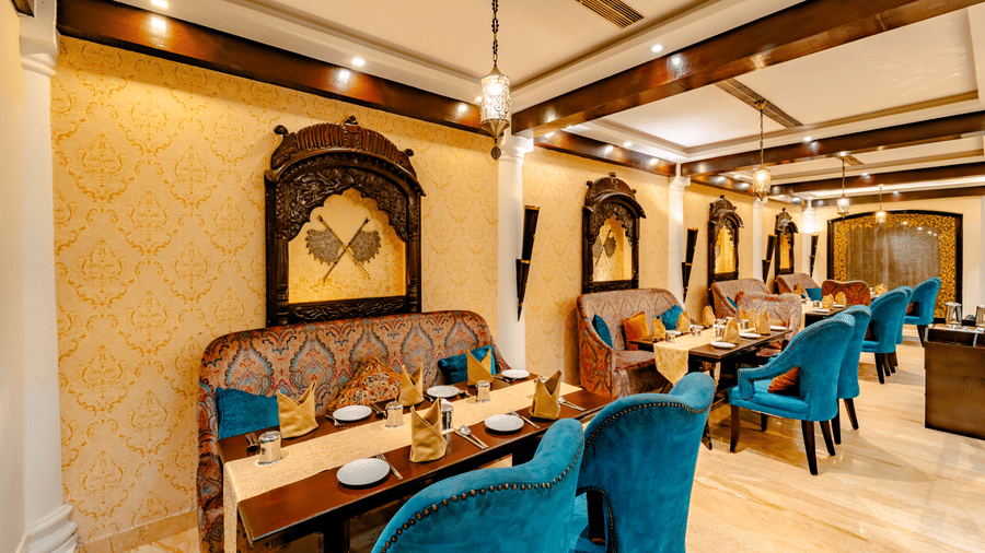 A sophisticated dining room at Za-aiqa, Noor-Us-Sabah Palace, Bhopal, featuring golden patterned wallpaper, ornate wooden wall hangings, and plush teal velvet seating arranged for a meal.