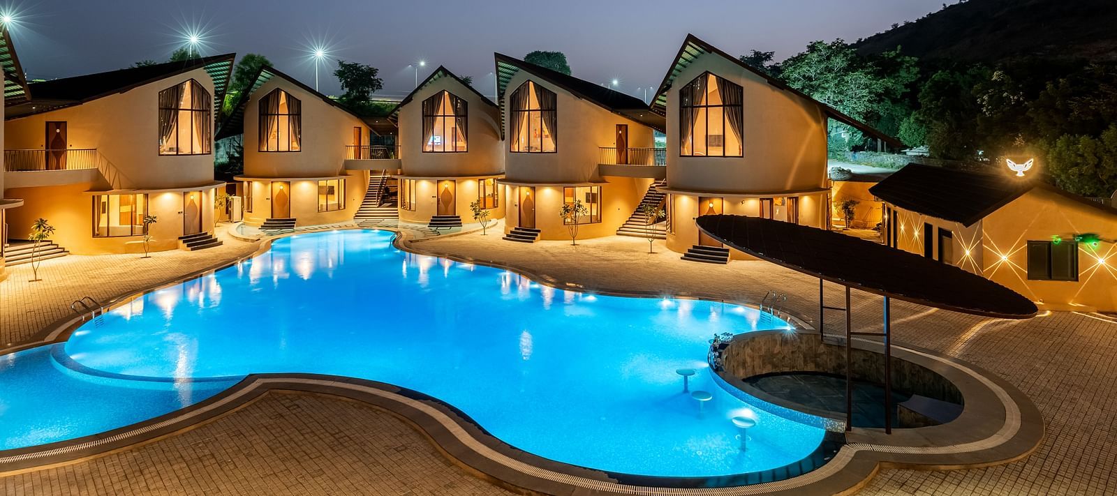 The Qualia Resort Club & Brewery, Udaipur shaped like a bean, surrounded by buildings with sloped roofs, set against a backdrop of trees and twilight sky.