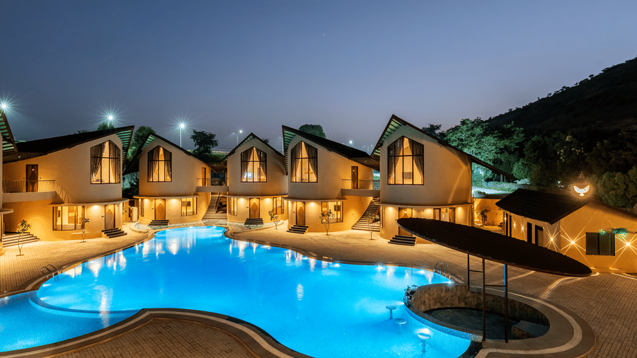 The Qualia Resort Club & Brewery, Udaipur shaped like a bean, surrounded by buildings with sloped roofs, set against a backdrop of trees and twilight sky.