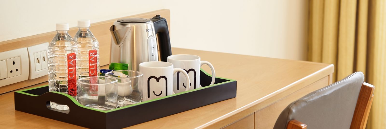 A close-up of a well-lit hotel room desk at Monday Hotels Mahape, Navi Mumbai, with a kettle and a tray of beverages.