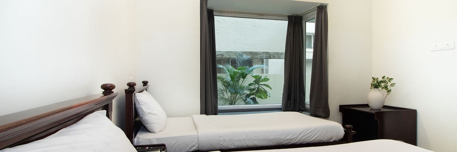 A bright hotel room featuring two twin beds with white linens, wooden furniture, a glass window, and soft lighting for a relaxing atmosphere at Sanctum Suites Indiranagar, Bangalore.