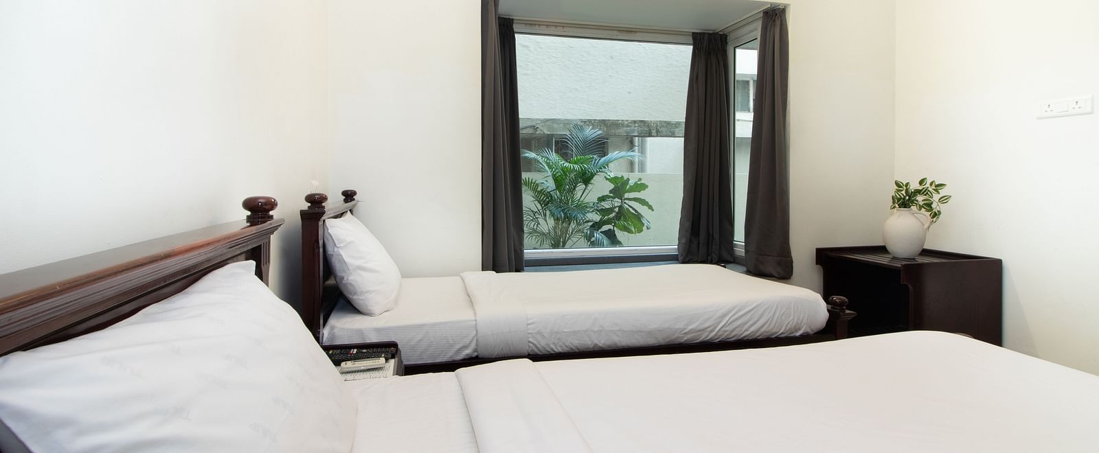 A bright hotel room featuring two twin beds with white linens, wooden furniture, a glass window, and soft lighting for a relaxing atmosphere at Sanctum Suites Indiranagar, Bangalore.