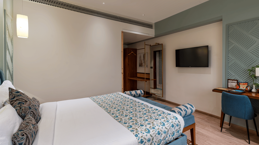 Side view of the Premium Room showing a neatly made double bed with blue bed runners, a wall-mounted television, with the enterace passage beside it - Amantra Comfort Hotel