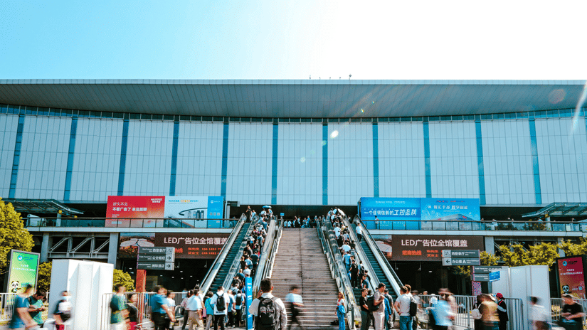 Crowds of people walking towards a large, modern exhibition centre with escalators and blue banners under a clear sky.