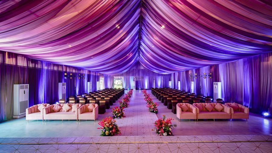 A banquet hall with decorated roof in purple colour and seating areas with flower pots along the aisle at Grande Bay Resort & Spa.