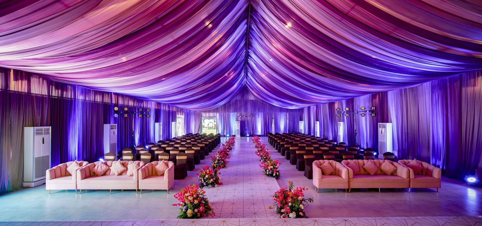A banquet hall with decorated roof in purple colour and seating areas with flower pots along the aisle at Grande Bay Resort & Spa.