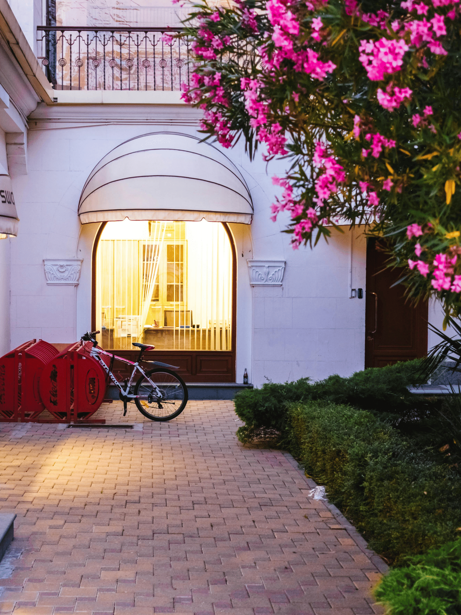 The exterior of a boutique shop with white awnings, a bike and a red cart outside, and vibrant pink flowering bushes.