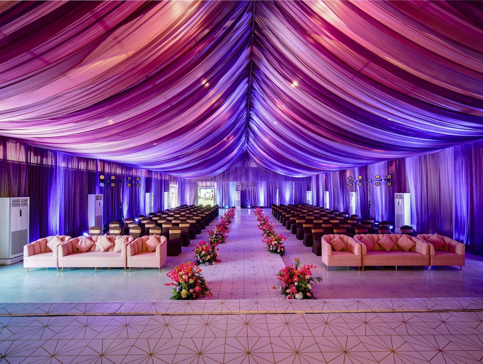 A banquet hall with decorated roof in purple colour and seating areas with flower pots along the aisle at Grande Bay Resort & Spa.