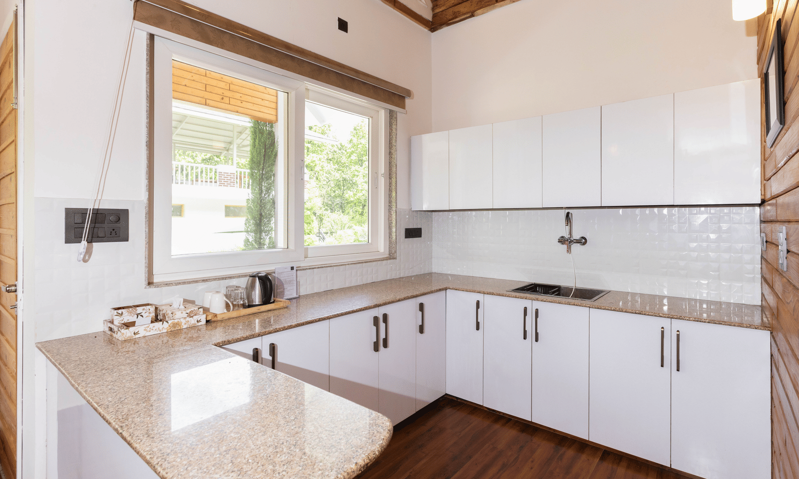 A white kitchen with wooden flooring, a stove, and large windows offering a view is visible - Megha Resort & Villas, Dehradun