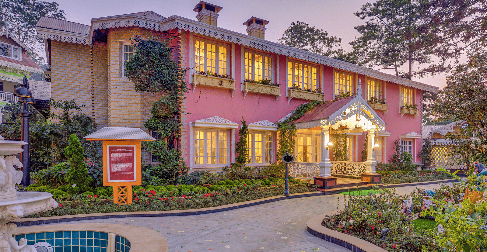 The exterior of the heritage-style MAYFAIR Himalayan Spa Resort Kalimpong with lit windows, a stone path, and a courtyard fountain.