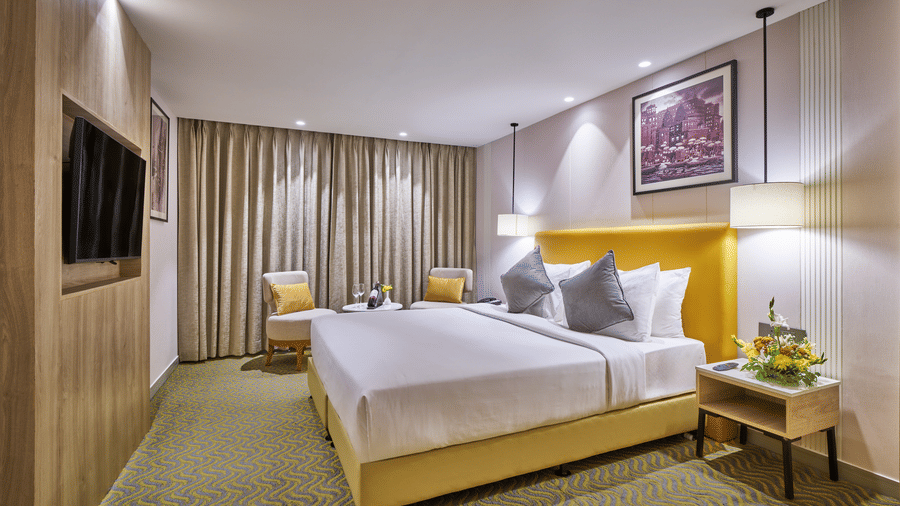 A guest room with a double bed, yellow headboard, bedside lamp, and a seating area with 2 armchairs | HHI Varanasi