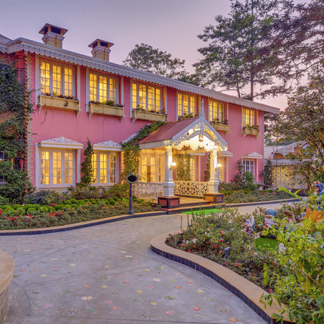Exterior view of the pink colonial-style building at MAYFAIR Himalayan Spa Resort Kalimpong, featuring warm window lighting and a stone-paved walkway at dusk.