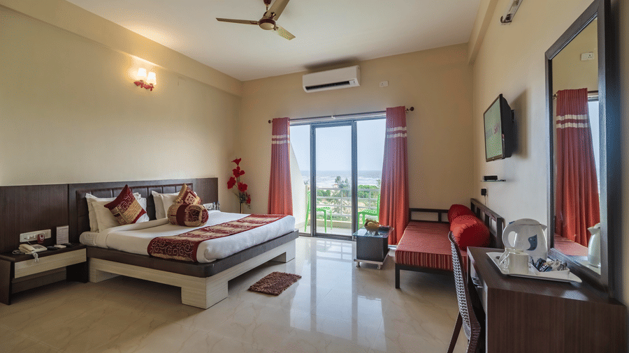 Modern room with king bed, red bench seating, desk, TV, and balcony overlooking greenery at Viceroy Beach & Spa Resort, Mandarmani