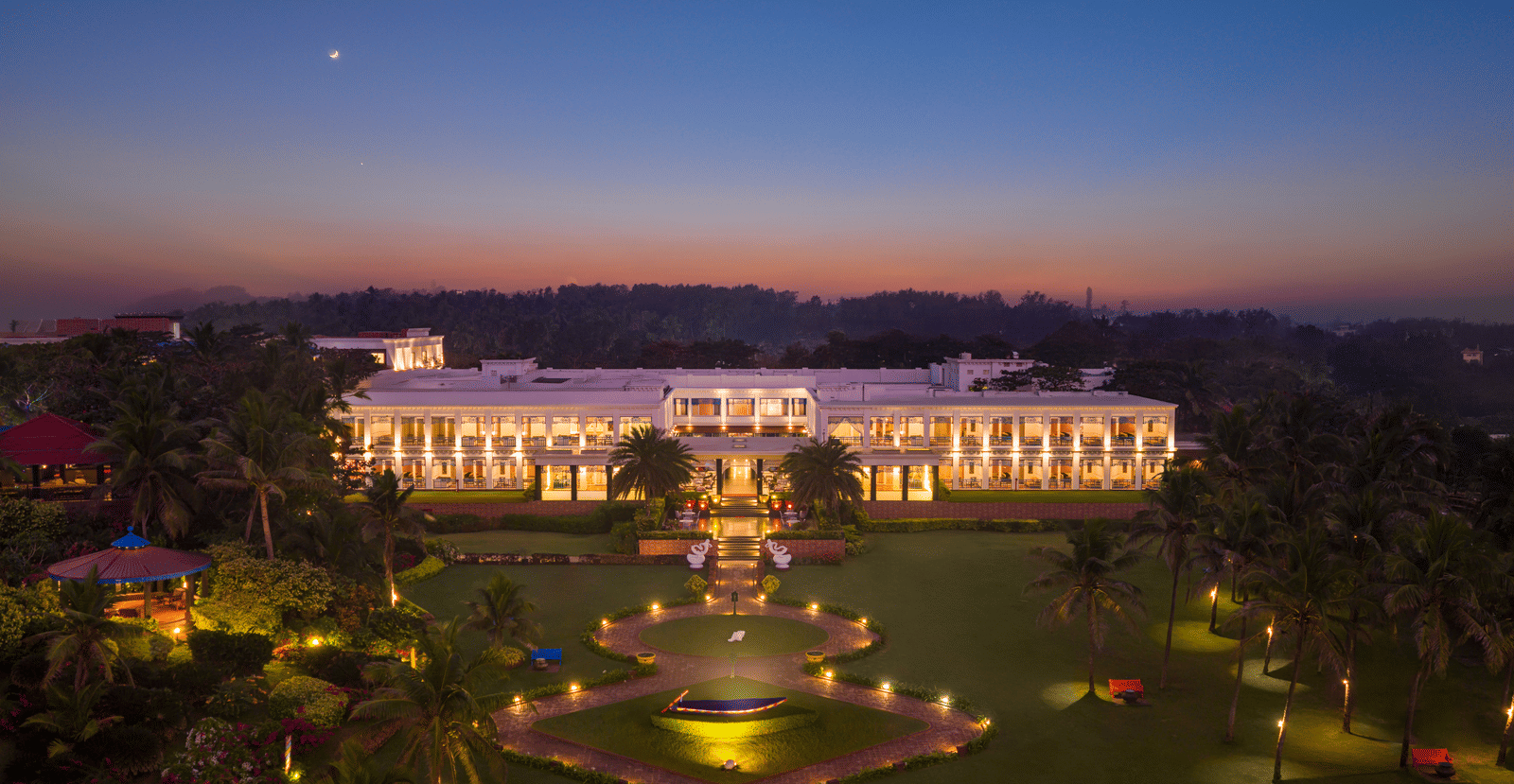 Aerial view of MAYFAIR Palm Beach Resort Gopalpur-On-Sea featuring the illuminated facade of the property and manicured lawns against an evening sky. 