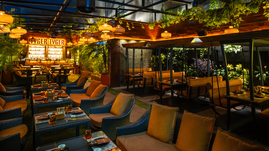 A restaurant seating space with long cushioned sofas, tables and glowing 'TYGER TYGER' sign at Infiniti Hotel, Indore.