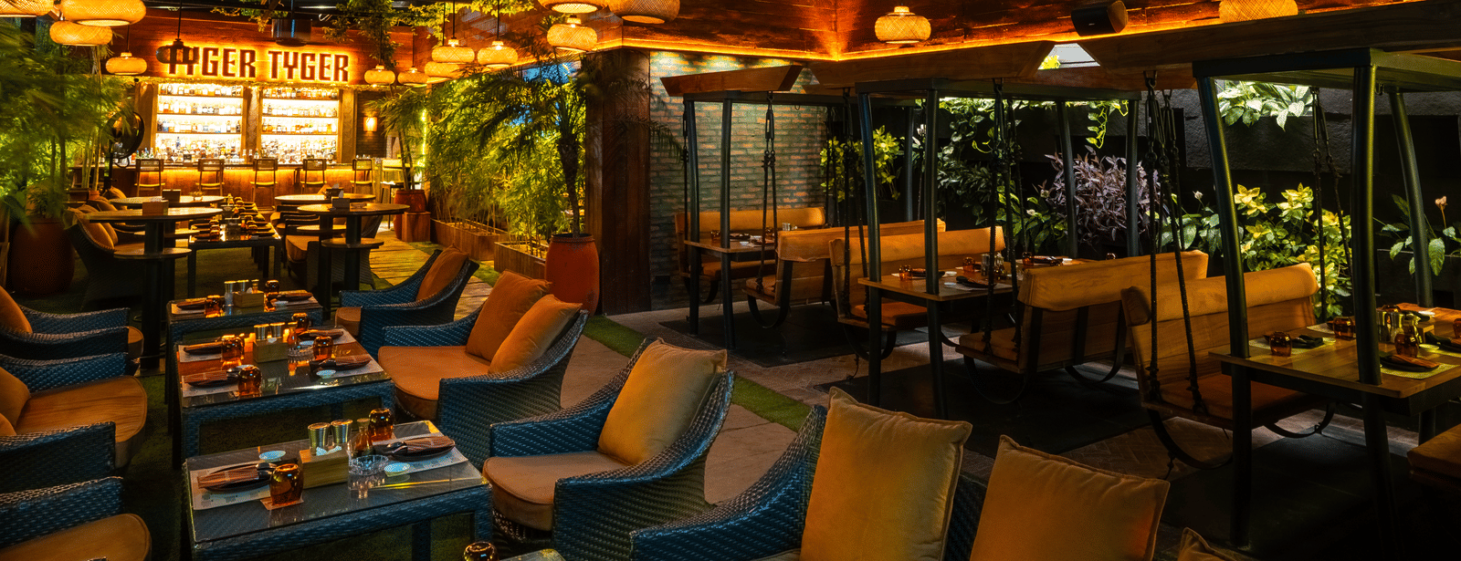 A restaurant seating space with long cushioned sofas, tables and glowing 'TYGER TYGER' sign at Infiniti Hotel, Indore.