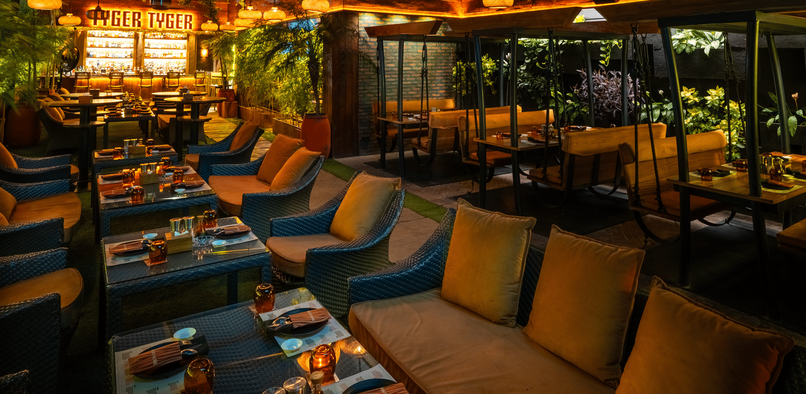 A restaurant seating space with long cushioned sofas, tables and glowing 'TYGER TYGER' sign at Infiniti Hotel, Indore.