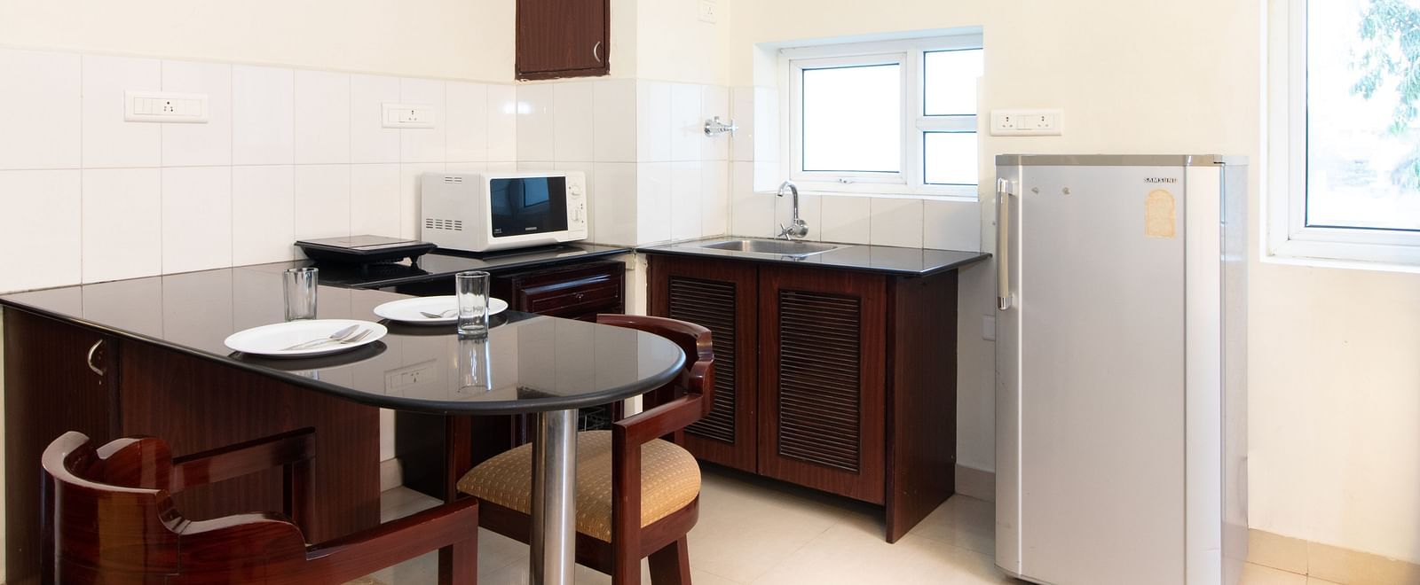 A compact kitchenette area with a dining table and chairs, a refrigerator, and a window for natural light at Sanctum Suites Indiranagar, Bangalore.