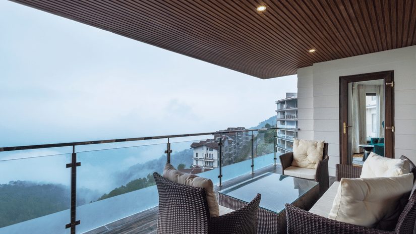 Private balcony with comfortable outdoor seating, glass railings, and panoramic views of Shimla’s misty hills and lush landscape, emphasizing serene relaxation spaces at The Manor Sports & Wellness Hotel.
