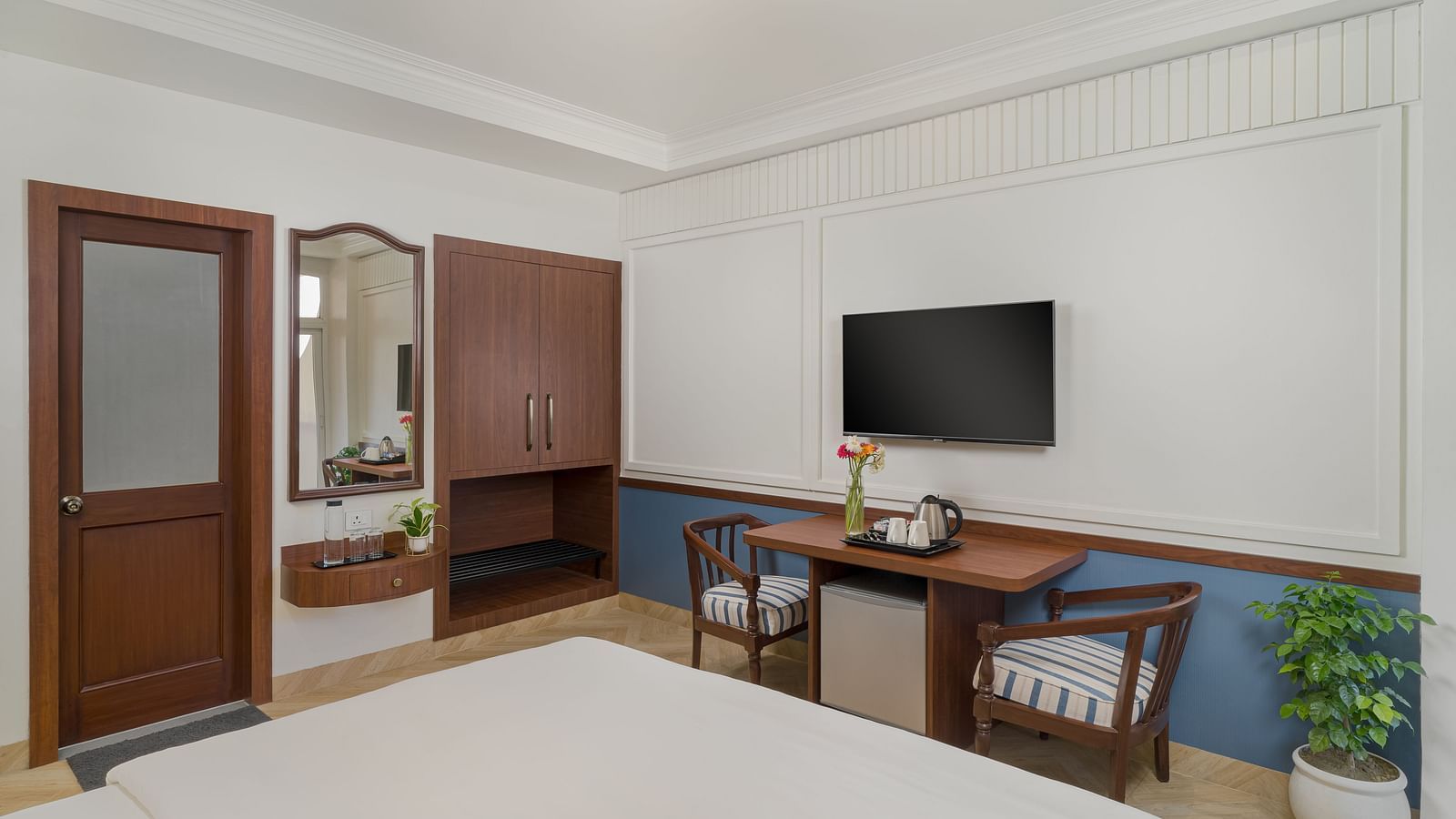 A room featuring a spacious bed, a wall-mounted television, a wooden table with a pair of wooden chairs beside it | Hotel Ganga Maiya