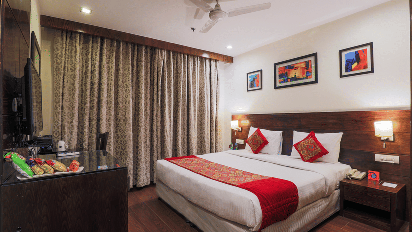 A hotel room showing a large bed with a red runner, a ceiling fan, snacks kept on a table opposite to the bed and colourful art works placed on the wall - Hotel Bluestone Prime (Nehru Place), New Delhi