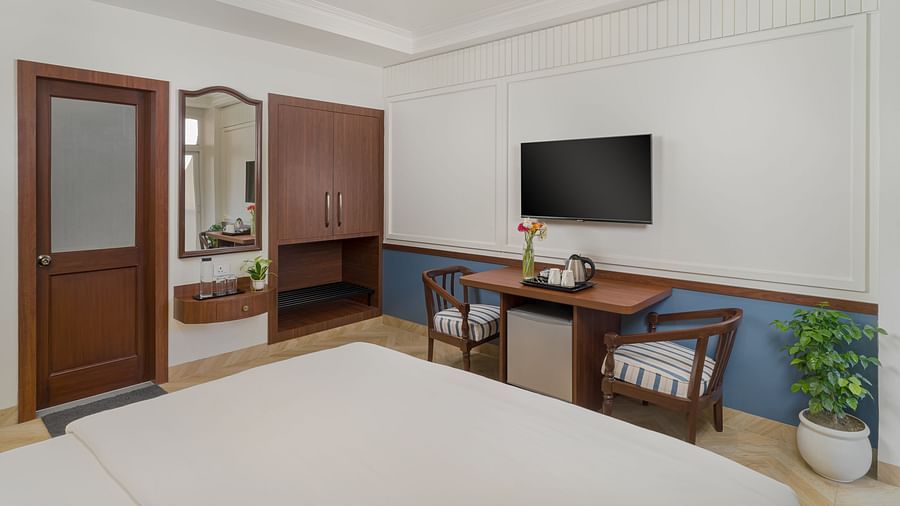 A room featuring a spacious bed, a wall-mounted television, a wooden table with a pair of wooden chairs beside it | Hotel Ganga Maiya