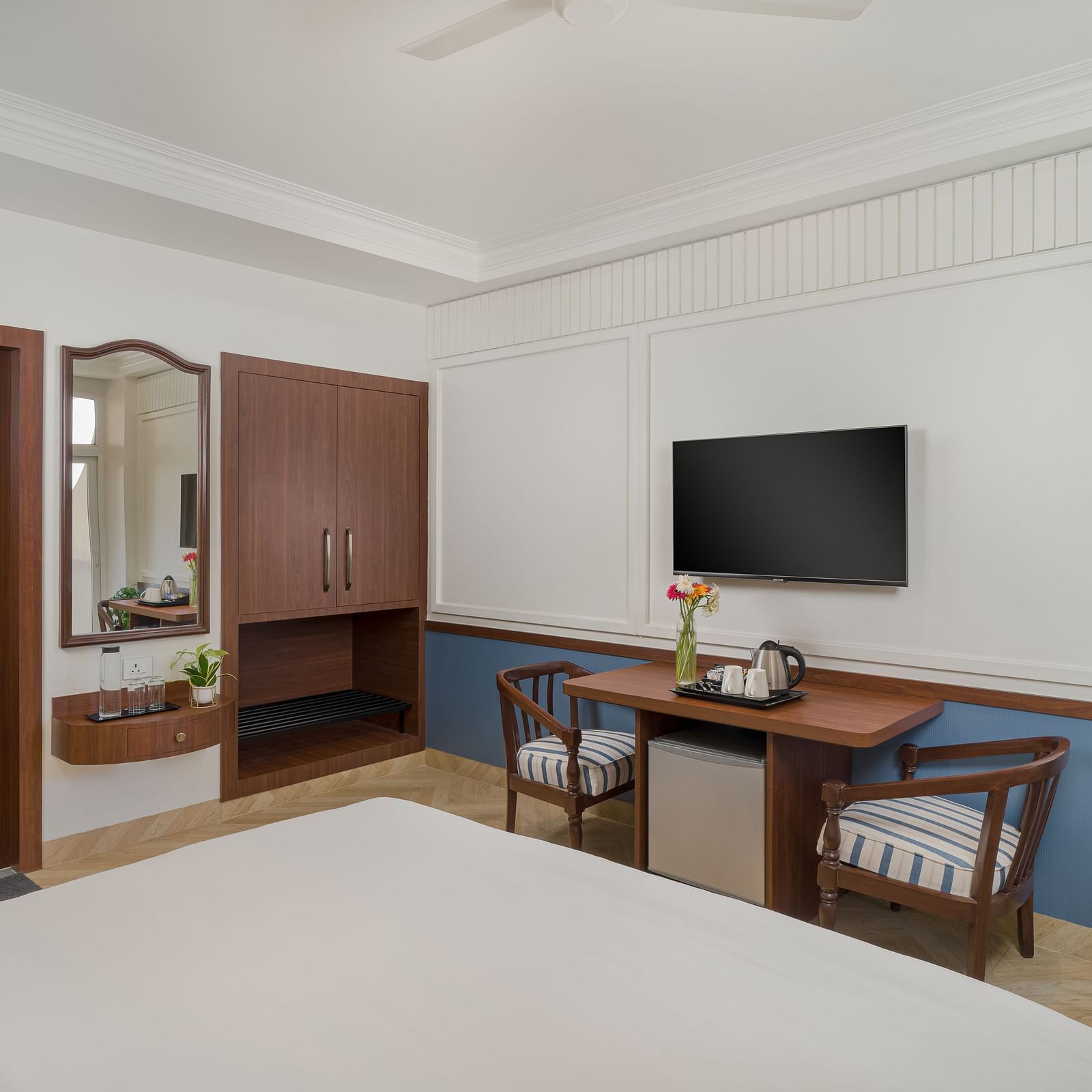 A room featuring a spacious bed, a wall-mounted television, a wooden table with a pair of wooden chairs beside it | Hotel Ganga Maiya