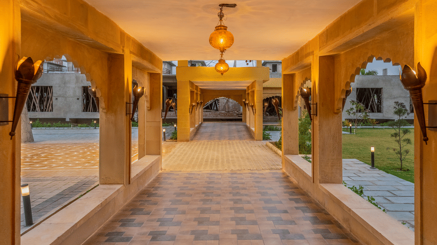 Stone corridor at EsthereaRaj Leela, Ranakpur featuring arched openings, wall-mounted torch lights, and hanging lanterns overlooking a garden.