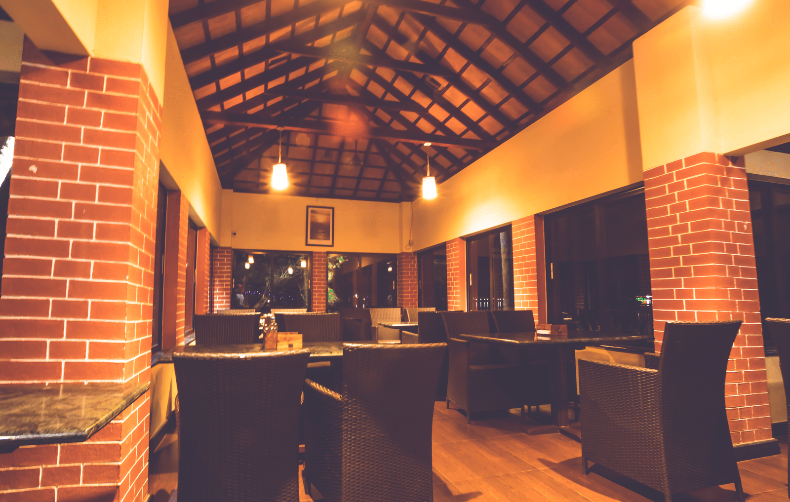 Dining space with wooden tables and chairs, brick pillars, and sloped wooden ceiling with lights at Paradise Lagoon Resort, Udupi.