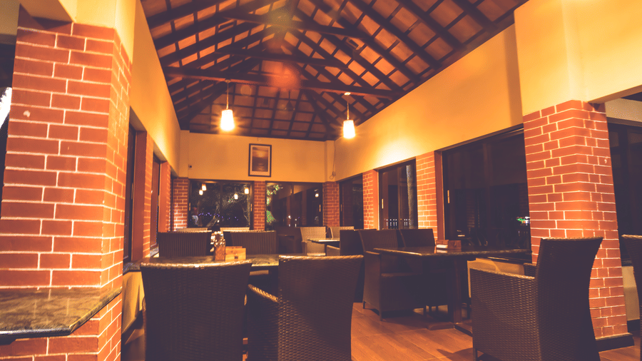 Dining space with wooden tables and chairs, brick pillars, and sloped wooden ceiling with lights at Paradise Lagoon Resort, Udupi.
