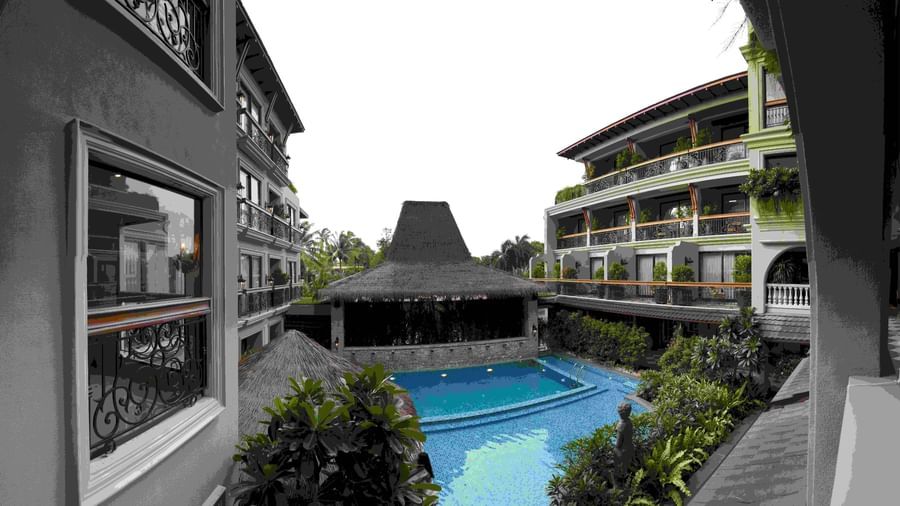 The outdoor swimming pool area at The Evren, Vagator, with a resort-like feel. The pool is surrounded by lush greenery and shaded by thatched-roof structures. The two-story hotel buildings with balconies are visible on either side, creating a courtyard eff