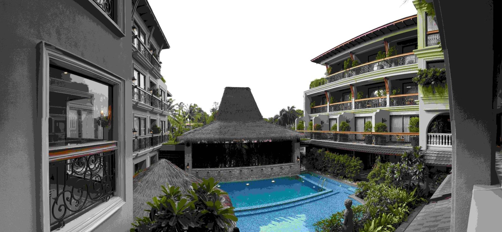 The outdoor swimming pool area at The Evren, Vagator, with a resort-like feel. The pool is surrounded by lush greenery and shaded by thatched-roof structures. The two-story hotel buildings with balconies are visible on either side, creating a courtyard eff