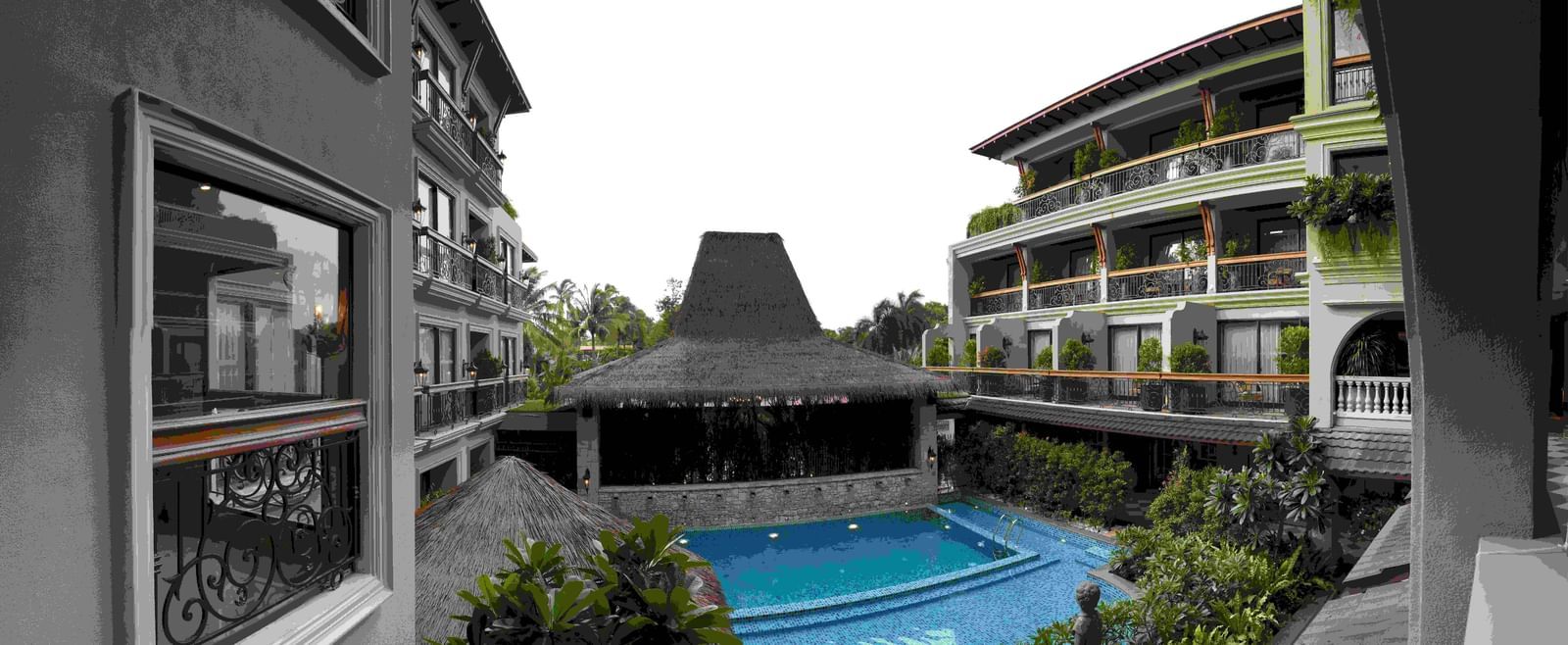 The outdoor swimming pool area at The Evren, Vagator, with a resort-like feel. The pool is surrounded by lush greenery and shaded by thatched-roof structures. The two-story hotel buildings with balconies are visible on either side, creating a courtyard eff