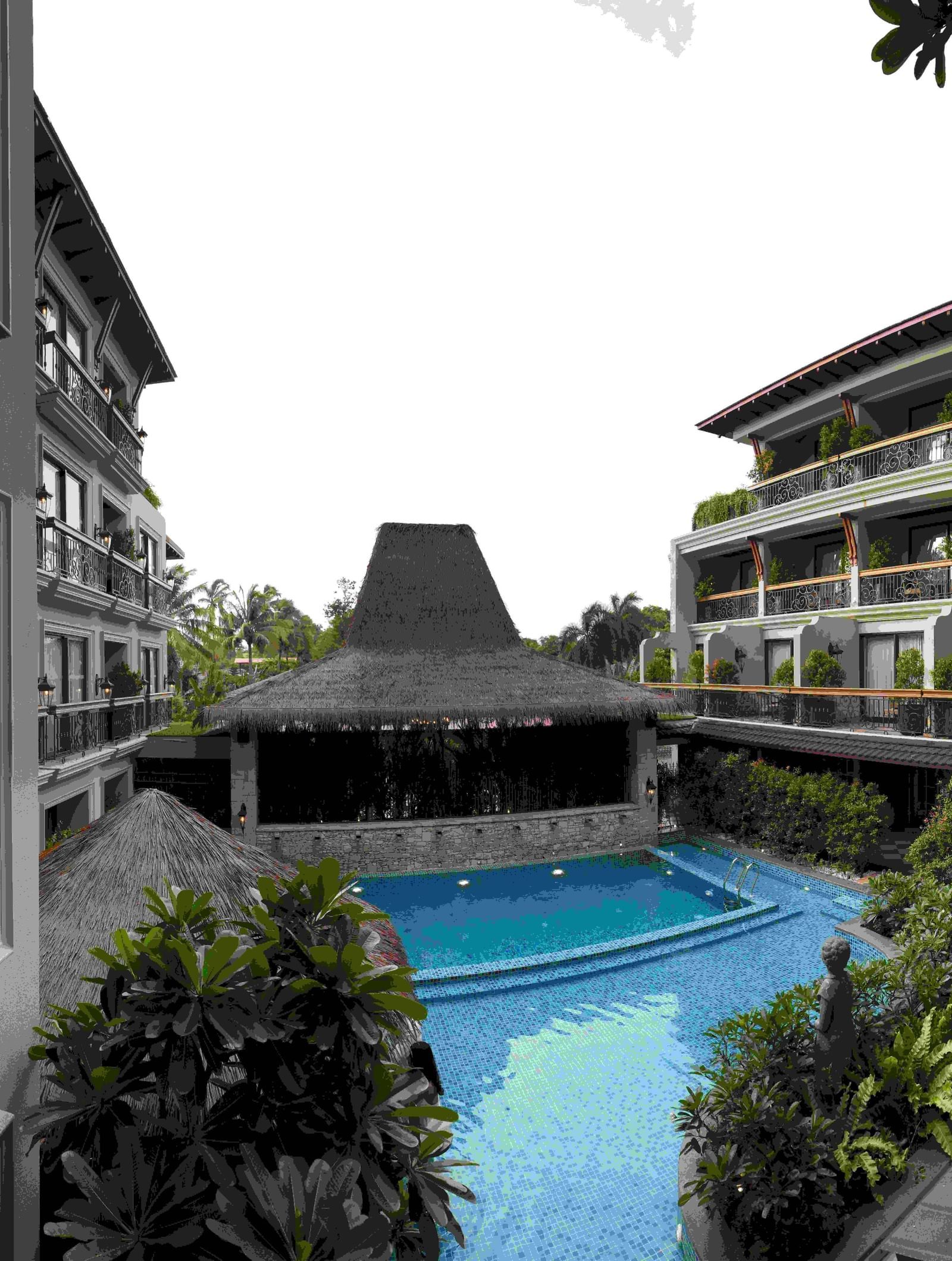 The outdoor swimming pool area at The Evren, Vagator, with a resort-like feel. The pool is surrounded by lush greenery and shaded by thatched-roof structures. The two-story hotel buildings with balconies are visible on either side, creating a courtyard eff