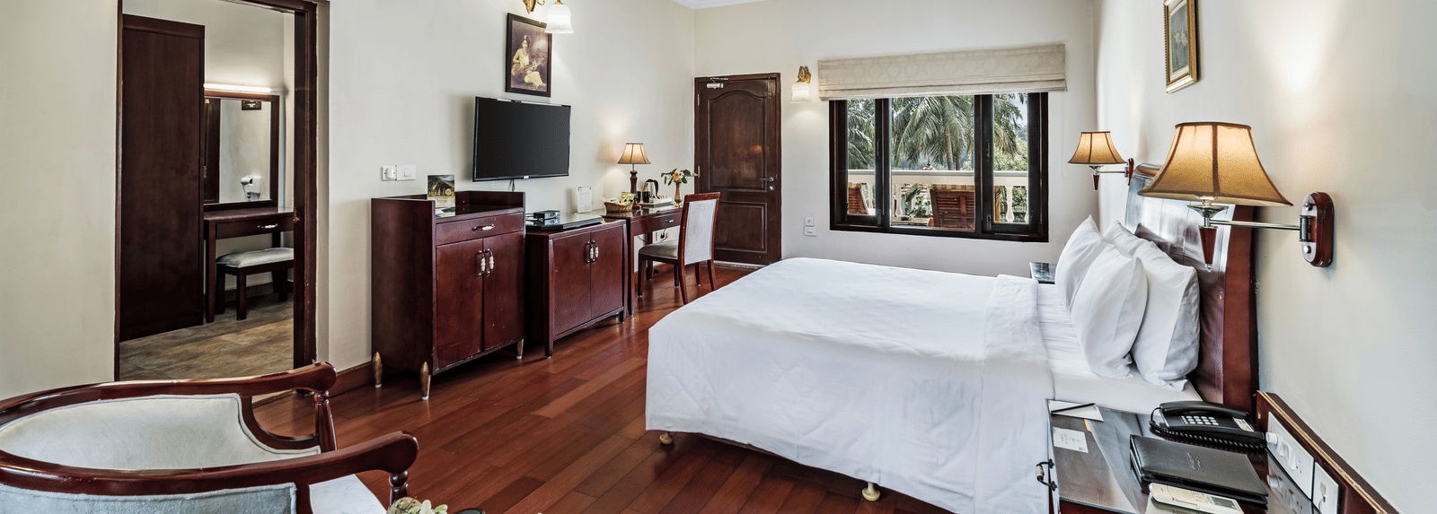 An image of the Garden room with river view featuring a queen size bed, bed side tables, lamps, ceiling fan, flat screen TV, and chairs - GReaT Trails River View Resort Thanjavur By GRT Hotels