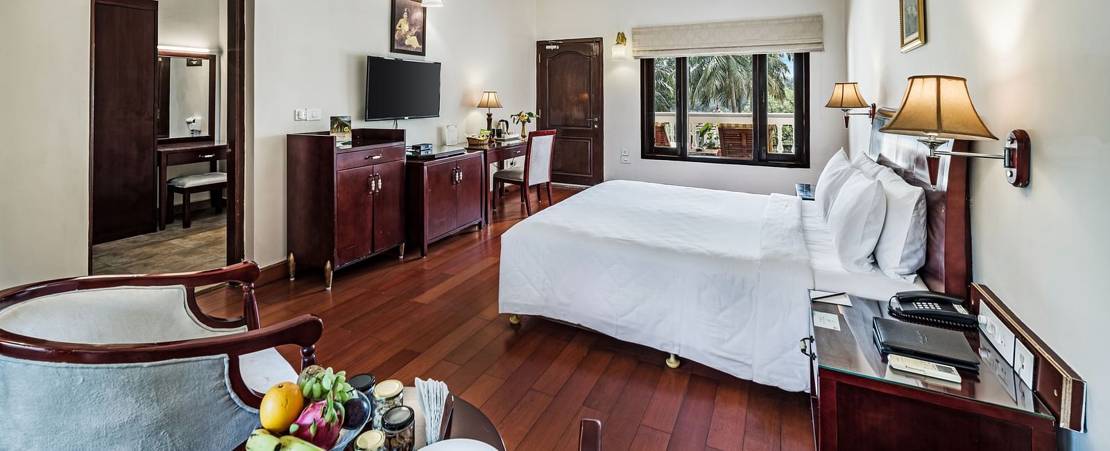 An image of the Garden room with river view featuring a queen size bed, bed side tables, lamps, ceiling fan, flat screen TV, and chairs - GReaT Trails River View Resort Thanjavur By GRT Hotels