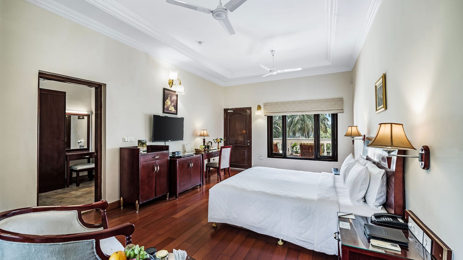 An image of the Garden room with river view featuring a queen size bed, bed side tables, lamps, ceiling fan, flat screen TV, and chairs - GReaT Trails River View Resort Thanjavur By GRT Hotels