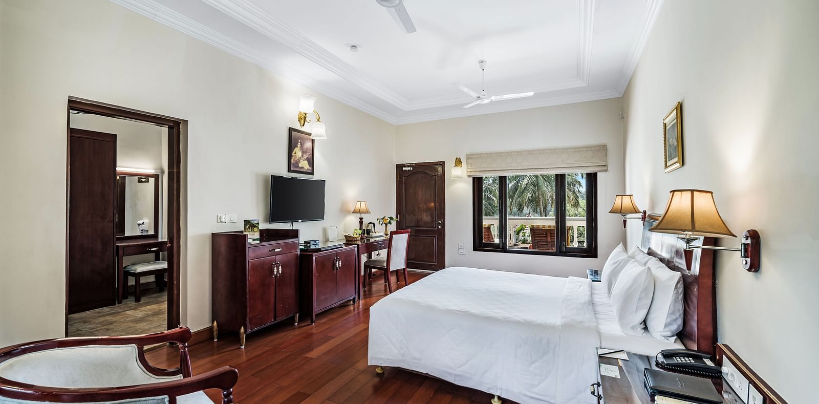 An image of the Garden room with river view featuring a queen size bed, bed side tables, lamps, ceiling fan, flat screen TV, and chairs - GReaT Trails River View Resort Thanjavur By GRT Hotels