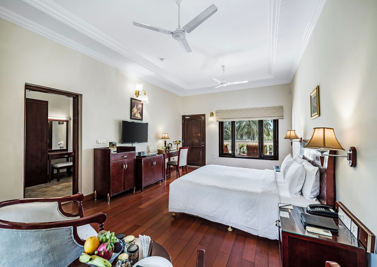 An image of the Garden room with river view featuring a queen size bed, bed side tables, lamps, ceiling fan, flat screen TV, and chairs - GReaT Trails River View Resort Thanjavur By GRT Hotels
