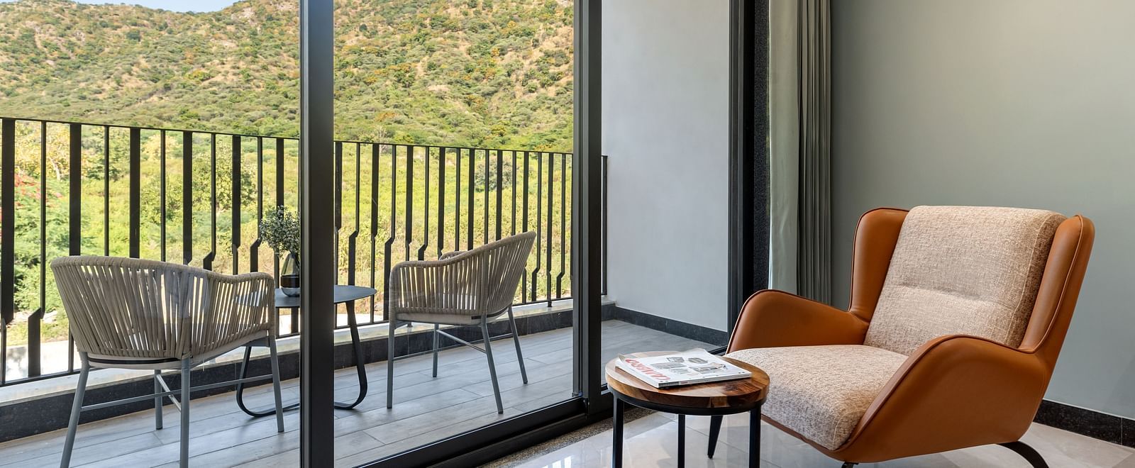Club Suite with Valley View Sitout with a balcony with outdoor seating, offering views of mountains and greenery through large glass doors at The Qualia Resort Club & Brewery, Udaipur.