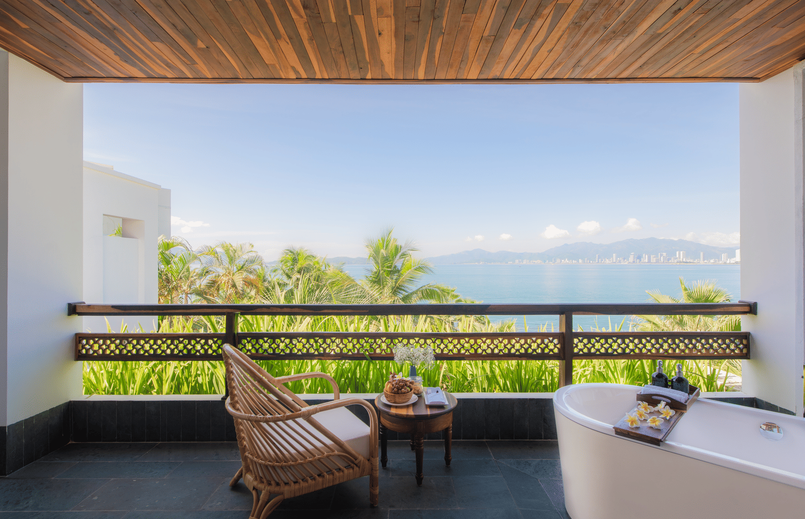Ocean View Premium balcony at Alibu Resort Nha Trang, Vietnam with bathtub, chair, table, glass railing, potted plants, facing water view and open sky, high ceiling, wide horizontal perspective.